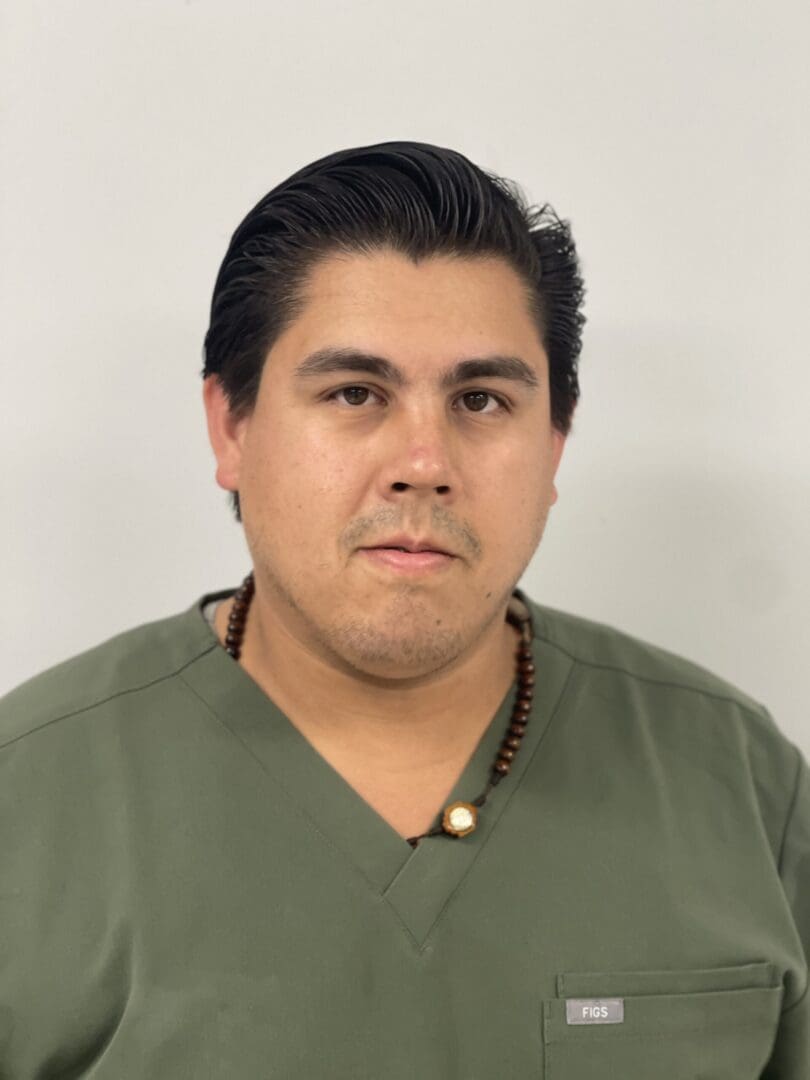 A man in green shirt and necklace standing next to wall.